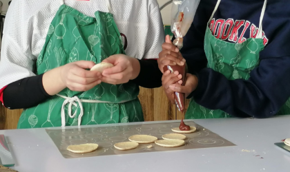 A gauche on voit une adolescente qui tient entre ses mains la croute d'un macaron tandis qu'à droite une autre adolescente presse une poche à douille remplie de chocolat sur des croutes de macarons posées sur un tapis de cuisson.