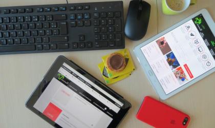Un bureau sur lequel sont posés divers éléments : deux tablettes, un smartphone, un clavier de PC, une souris, une tasse de café et un biscuit."