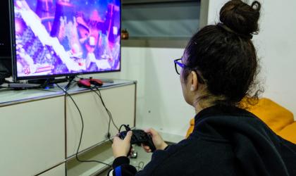 Accéder à l'événement "Jeu vidéo : FIFA 2022 sur PS4". Visuel : A droite, de dos est assise une jeune personne portant un chignon haut, des lunettes et un gilet noir. Elle tient à la main une manette connecté à un écran situé à gauche de la photo et sur lequel est affiché une partie de jeu vidéo.