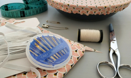 Accédez à l'événement "Atelier couture". Visuel : Sur une table sont éparpillés une paire de ciseaux, un étui à aiguilles, un rouleau de fil blanc, un mètre-ruban, du tissu à motifs roses et des épingles à nourrice.