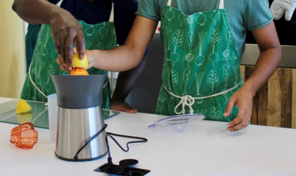 Accédez à l'événement "Atelier cuisine : Jus santé". Visuel : Deux personnes portant un tablier de cuisine sont debout devant un plan de travail et pressent un citron à l'aide d'un presse-agrume visible en premier plan.