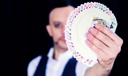 Un magicien tient face caméra, de sa main gauche, des cartes à jouer rangées en arc de cercle. Les cartes dissimules la moitiée gauche de son visage.
