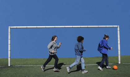 Trois femmes courent vers la droite, l'une derrière l'autre en passant devant une cage de but. La femme qui est en tête de file a un ballon de football orange à ses pieds.