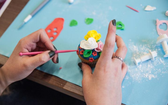 On voit les mains d'une personne qui tient un cupcake de sa main droite et avec sa main gauche tient un outil en forme de stylet permettant de former les fleurs en pâte à sucre sur le dessus du cupcake.