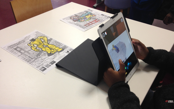 Une table blanche avec un coloriage représentant un robot géant jaune. Un enfant tient une tablette devant le coloriage, sur laquelle on peut voir l'image s'animer en 3D.