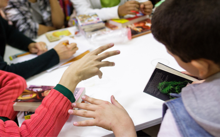 Un groupe est autour d'une table blanche. Des mangas se trouvent sur la table et une personne, de dos, en tient un à la main. Une autre personne écrit ou dessine sur une feuille.