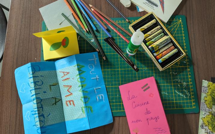 photo d'une table de travail ou sont disposé des crayons de couleur, pastels, un tube de colle et trois exemples de zines, un plié dont la couverture lit "la cuisine de mon pays" et un deplié qui lit "tout le monde aime la bibliothèque"